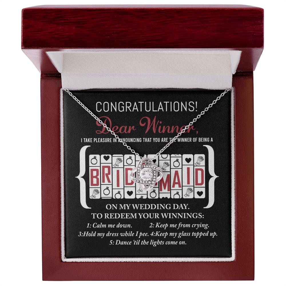 A "To Bridesmaid, The Winner Of" jewelry box in red and white, containing a Love Knot Necklace adorned with cubic zirconia crystals. Inside the lid, a card reads: "Congratulations! Dear Winner, I take pleasure in announcing that you are the winner of being a Bridesmaid on my wedding day. To redeem your winnings..." along with various tasks.