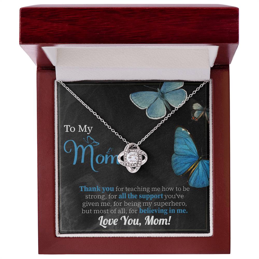 The "To Mom, Believing In Me - Love Knot Necklace" comes in a box adorned with cubic zirconia crystals and features a heartfelt message that reads "To My Mom," expressing deep gratitude and love, along with a charming butterfly illustration in the background.