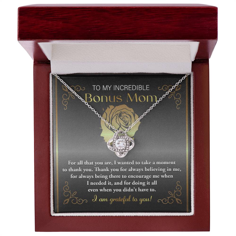 A stunning "To Bonus Mom, To Encourage Me - Love Knot Necklace" in silver, featuring intertwining cubic zirconia crystals in the pendant, is carefully placed inside a red box. The lid of the box bears a heartfelt message for a "Bonus Mom," expressing gratitude for her support and encouragement.