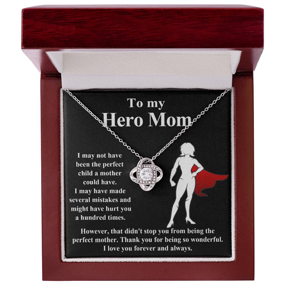 The "To Mom, The Perfect Mother - Love Knot Necklace" is showcased in an open red box. Inside, the box displays a heartfelt message of appreciation to a mother, reading "To my Hero Mom," accompanied by a silhouette of a woman in a cape. The necklace dazzles with cubic zirconia crystals and is available in your choice of white gold or yellow gold finish.