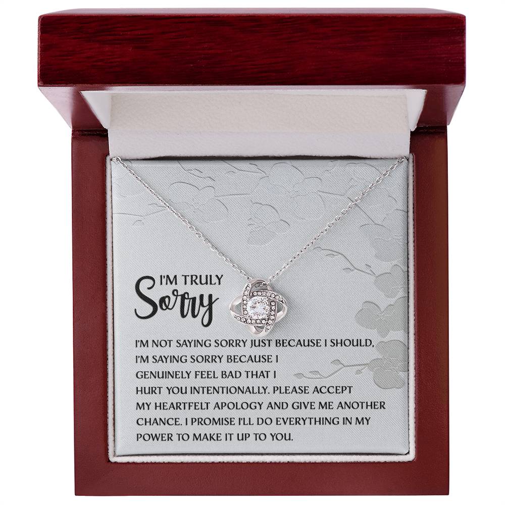 Introducing the Sorry-Feel Bad - Love Knot Necklace, featuring a pendant on a card with an apology message: "I'm truly sorry. I'm not saying sorry just because I should..." and continues with more heartfelt text. The necklace showcases a sparkling cubic zirconia crystal set in your choice of white gold or yellow gold finish, all against a light floral design background.
