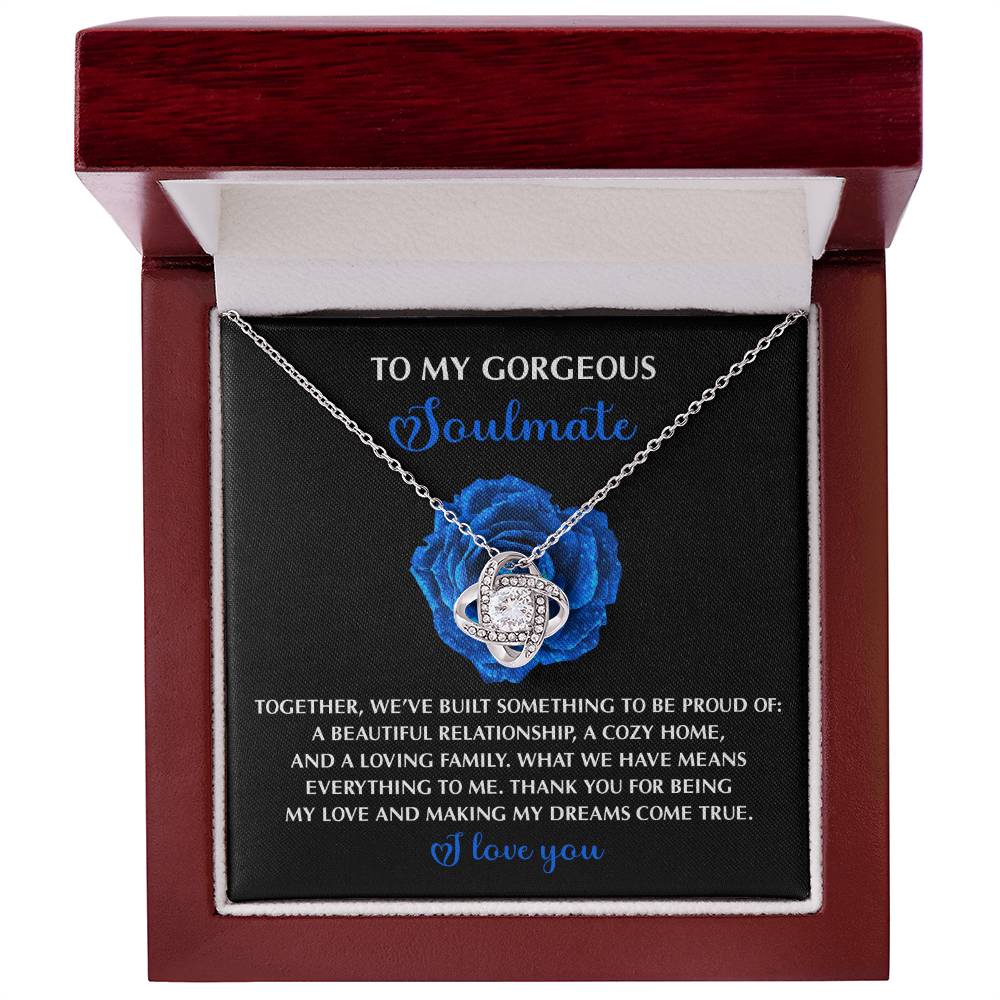 The Soulmate-Dreams Come True - Love Knot Necklace, featuring a gold finish and cubic zirconia crystals, is nestled in a box with a sentimental message on a black card, elegantly highlighted by a blue rose backdrop.