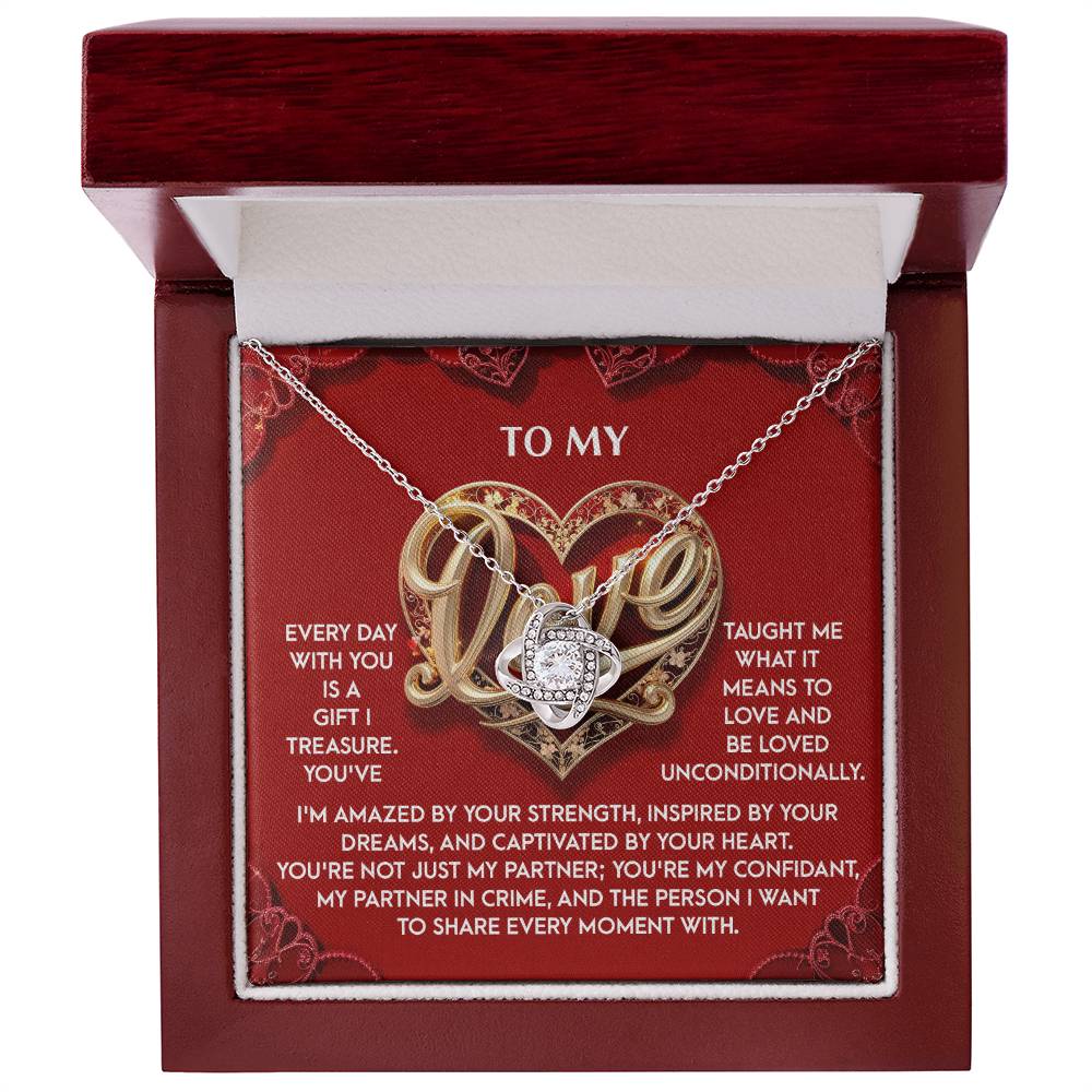 The "My Love-Every Moment - Love Knot Necklace" features a heart-shaped silver pendant with cubic zirconia crystals, presented in a red box. The box includes a romantic message about love and partnership, addressed "To My Love," with intricate background designs, ideal for a personalized gift.