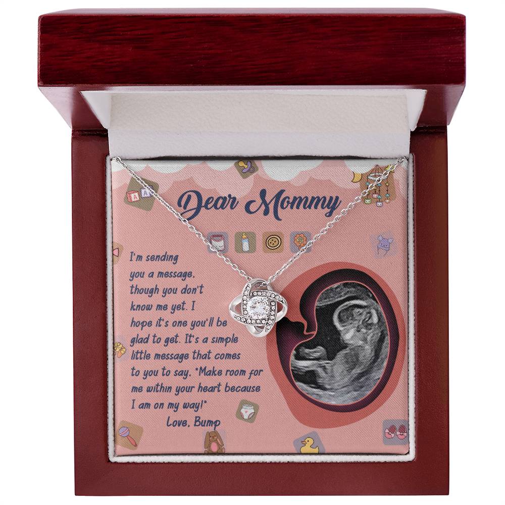 The "To Mom To Be, On My Way" Love Knot Necklace, adorned with cubic zirconia crystals, is elegantly displayed in an open red jewelry box. Accompanying it are a heartfelt message addressed to "Mommy" and an ultrasound image of a baby. The background features small decorative graphics, making this the perfect personalized gift.