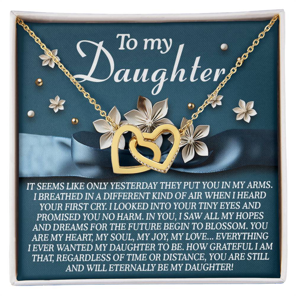 The Daughter-Hopes Dreams Interlocking Hearts Necklace, featuring gold and cubic zirconia, is set against a floral backdrop. The box lid says "To my Daughter" with a touching message about aspirations for her below this ideal gift.