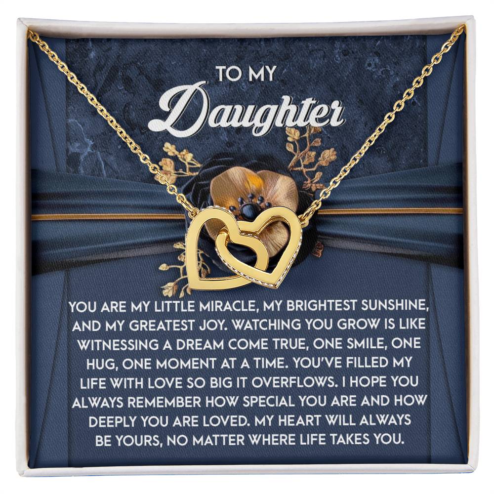 The Daughter-You Are Loved Interlocking Hearts Necklace, adorned with gold and cubic zirconia crystals, rests on an emotional message card for a daughter, set against a floral-accented dark blue background.