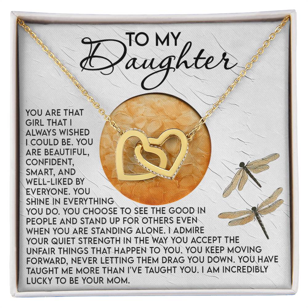 A decorative box with the Daughter-Quiet Strength - Interlocking Hearts Necklace on a yellow background. The necklace, embellished with cubic zirconia crystals, perfectly complements the heartfelt message to a daughter expressing love and admiration, featuring two dragonfly illustrations.