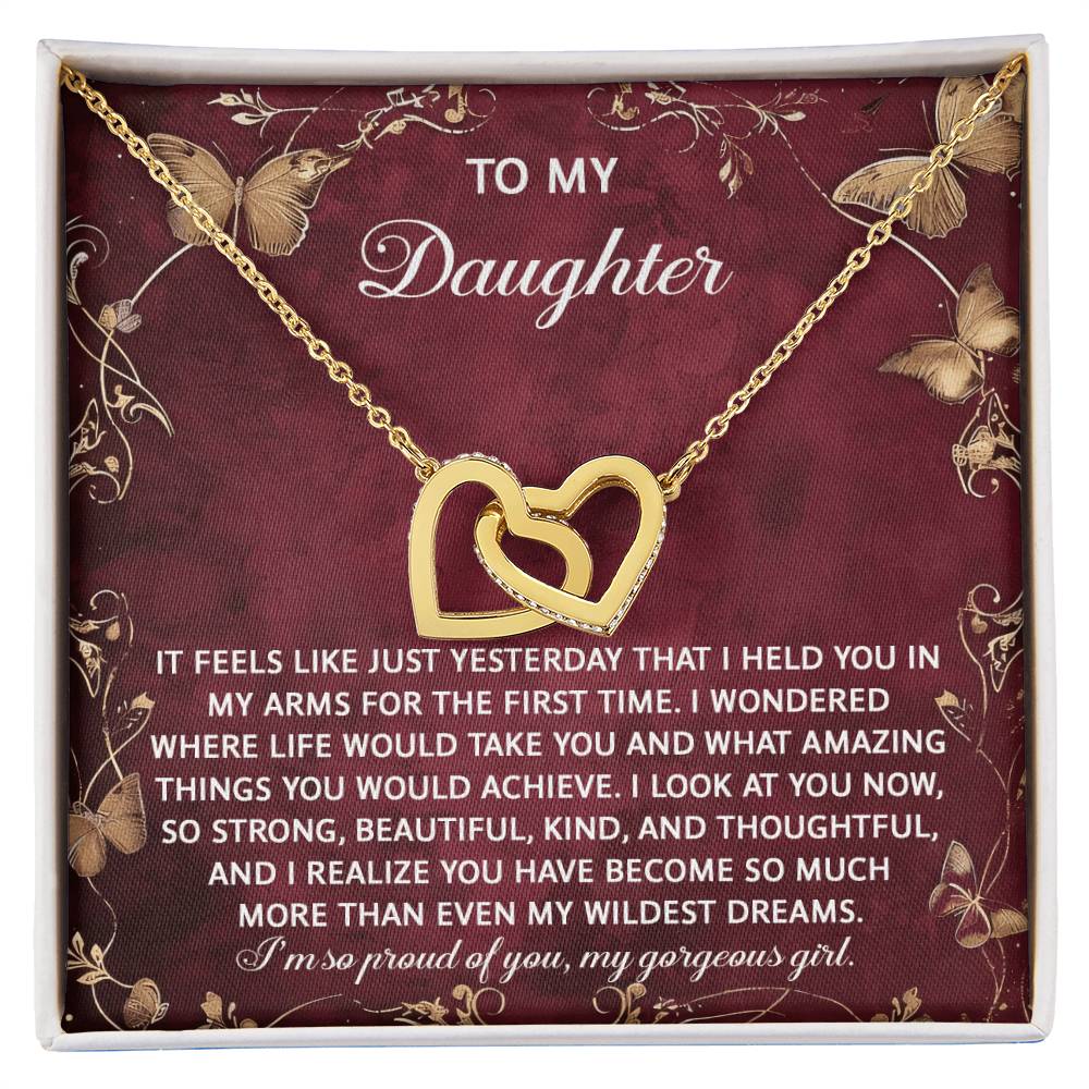 The Daughter-Wildest Dreams - Interlocking Hearts Necklace, featuring a rose gold finish and sparkling cubic zirconia crystals against a maroon backdrop, carries a heartfelt message for a beloved daughter.