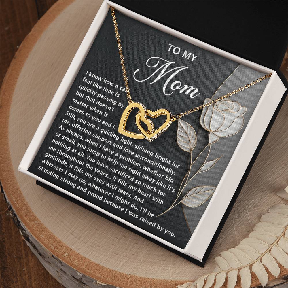 The Mom-Guiding Light Interlocking Hearts Necklace, featuring a rose gold finish and sparkling cubic zirconia crystals, sits on a "To My Mom" card with a heartfelt message in a gift box. The box is elegantly displayed on a wooden surface with a beige decorative cloth.