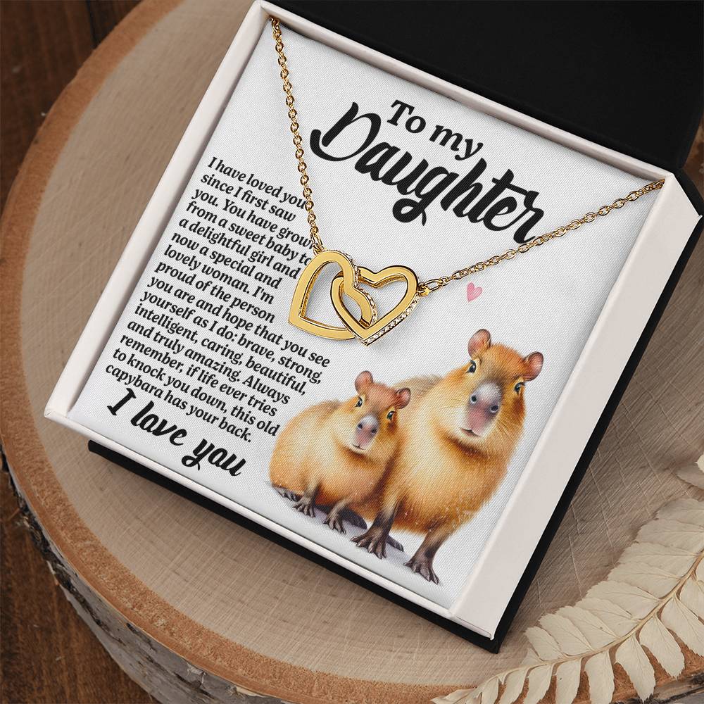 The Daughter-Sweet Baby - Interlocking Hearts Necklace, with cubic zirconia and a rose gold finish, rests on a card featuring two capybaras. It conveys love with the message "To my Daughter." The elegant box is displayed on a wooden table.