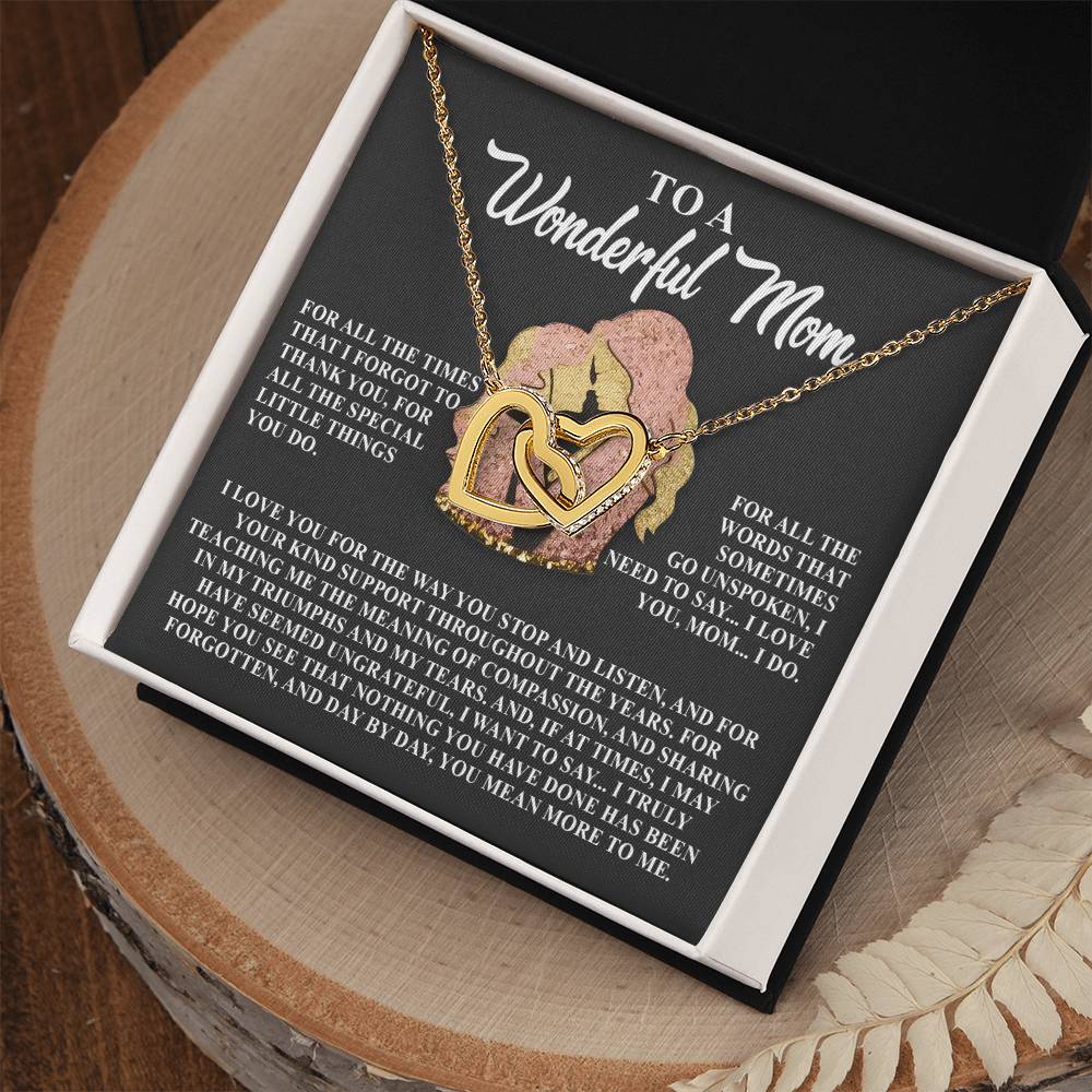 The Mom-More To Me - Interlocking Hearts Necklace features gold and rose gold intertwined hearts adorned with shimmering cubic zirconia. It rests on a black card inscribed with "To a Wonderful Mom" and a heartfelt message, all elegantly displayed in an open black box on a wooden surface.