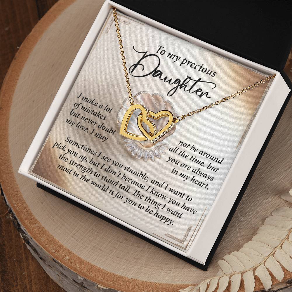 The "Daughter-Be Happy - Interlocking Hearts Necklace" features a sparkling cubic zirconia-adorned 18k yellow gold finish and rests in a jewelry box with a heartfelt message to your daughter, promoting love and encouragement. The box sits gracefully on a wooden surface.