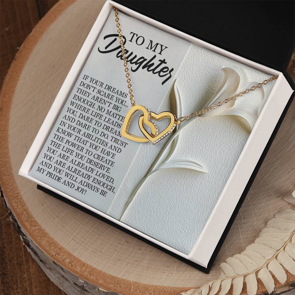 The Daughter-Dare Dream Interlocking Hearts Necklace in rose gold, adorned with sparkling cubic zirconia, rests on an open box alongside a heartfelt "my daughter" message. An embossed floral card complements the display on a circular wooden surface.