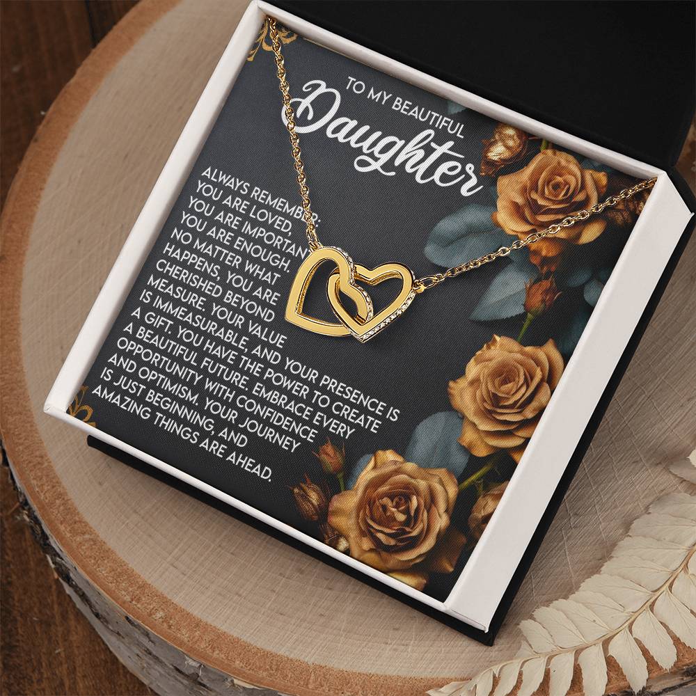 An open box showcases the Daughter-Loved Enough - Interlocking Hearts Necklace adorned with cubic zirconia on an orange rose background. The top reads "To My Beautiful Daughter," featuring a heartfelt message of love and encouragement. The box sits on a wooden surface with a feather decoration.