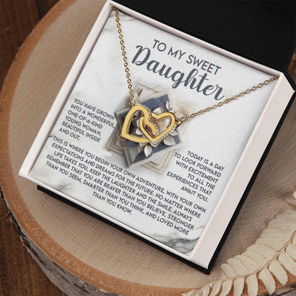 The Daughter-For The Future Interlocking Hearts Necklace, featuring heart pendants, is elegantly displayed on a card inside a black box. The card's message reads, "To my sweet daughter," with a loving note. Resting on a wooden surface, the box adds elegance to this thoughtful gift.