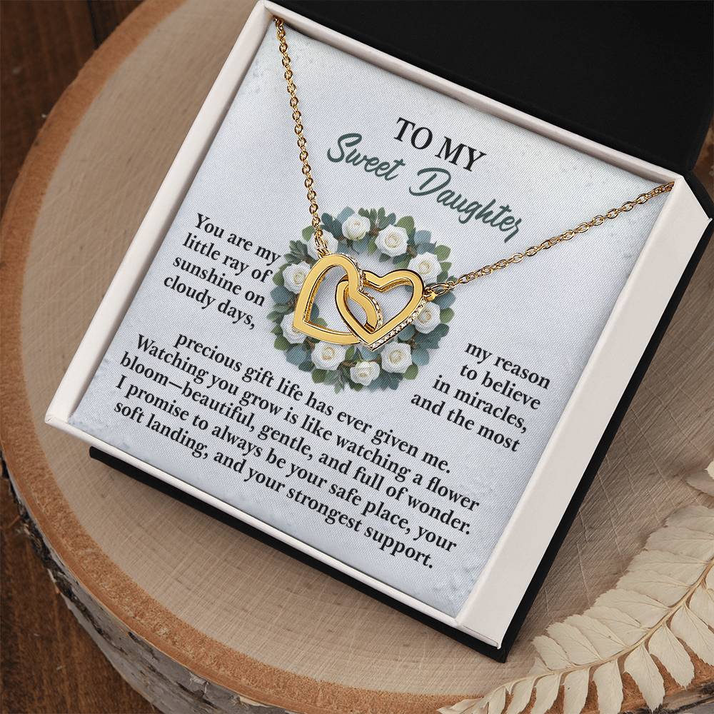 The Daughter-Strongest Support interlocking hearts necklace, embellished with cubic zirconia crystals, is presented in a black and white box. A heartfelt message to a daughter, adorned with white rose illustrations, appears in the background. The box sits on a wooden surface with decorative accents nearby.