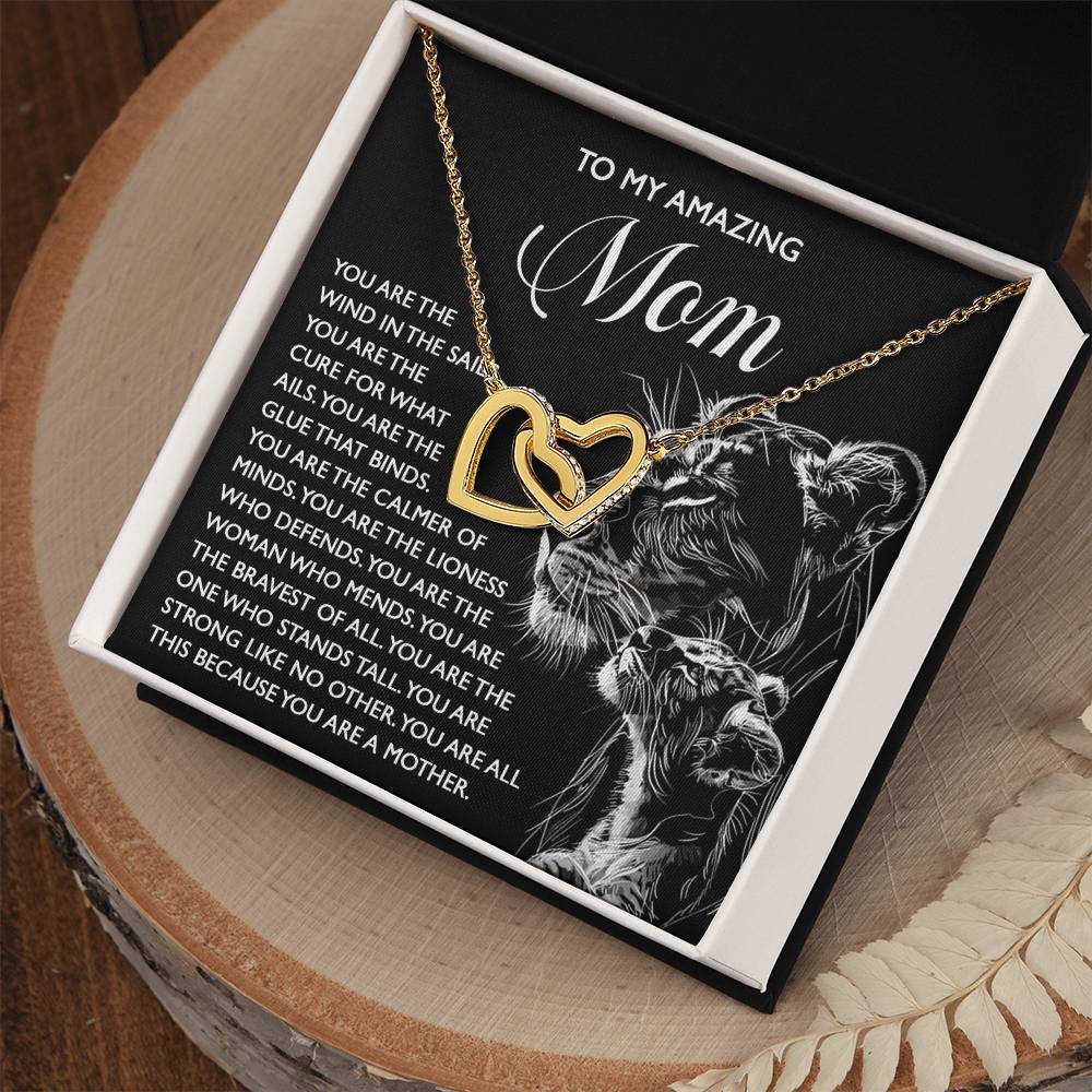 The gift box features the Mom-The Cure - Interlocking Hearts Necklace in rose gold with cubic zirconia. A card reads "To My Amazing Mom" next to a lioness illustration, while feather accents adorn the wooden surface.
