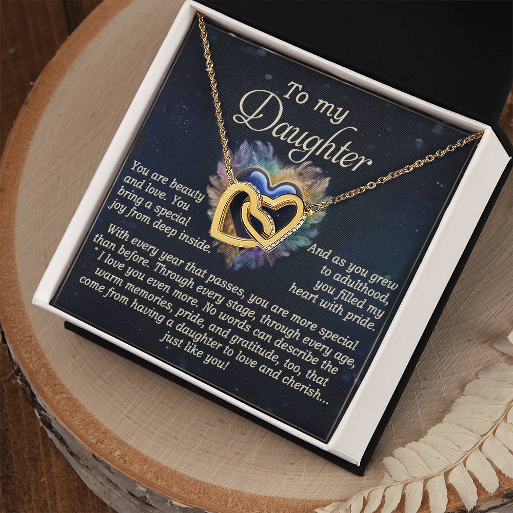 The "Daughter-Like You - Interlocking Hearts Necklace" features a gift box containing a necklace crafted from surgical steel with a rose gold finish and adorned with cubic zirconia crystals. It comes with a heartfelt message for a daughter, expressing love and admiration, all displayed on a wooden surface.