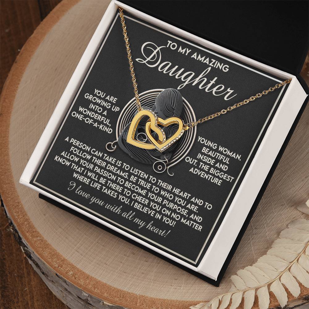 The Daughter-Your Dreams - Interlocking Hearts Necklace, adorned with sparkling cubic zirconia crystals and displayed in a black box, features a card reading, "To my amazing daughter," with an inspirational message about growth, passion, and belief. The box is elegantly set on a wooden surface.