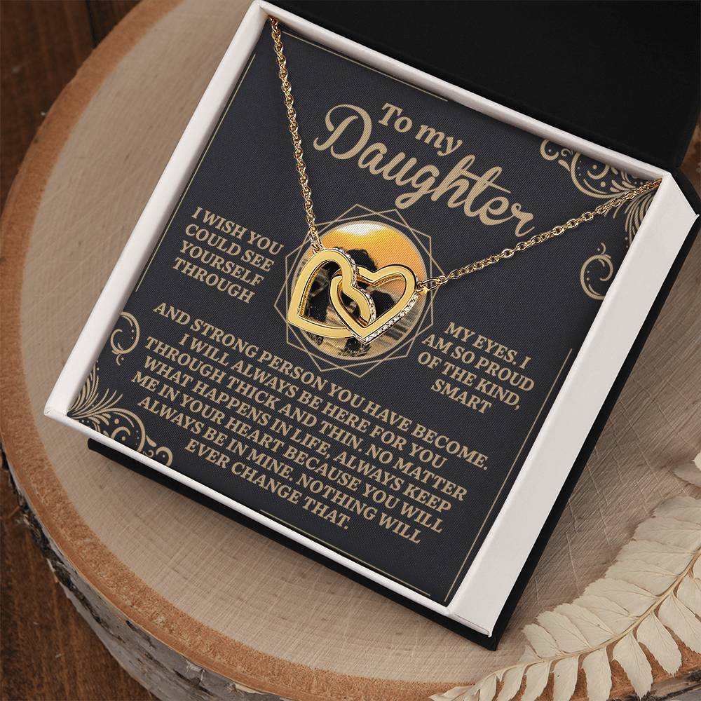 The Daughter-Proud Heart Interlocking Hearts Necklace, with a rose gold finish and cubic zirconia crystals, is elegantly presented in an open black box. The lid features an inspirational message for a daughter, expressing love and pride. The box rests beautifully on a wooden surface.