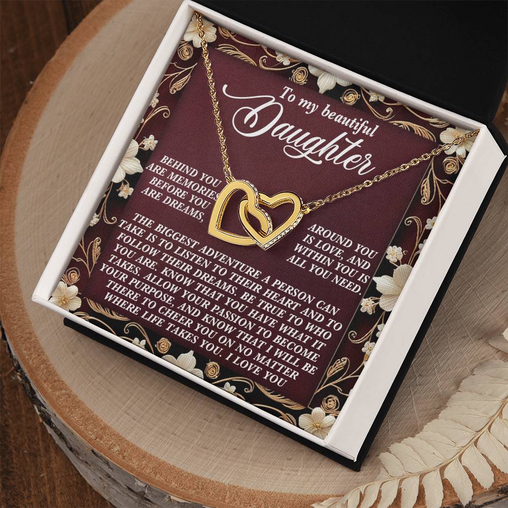 The Daughter-Follow Dreams - Interlocking Hearts Necklace is presented in an open box with a heartfelt message to a daughter. The background features floral decorations, and the box sits elegantly on a wooden surface.