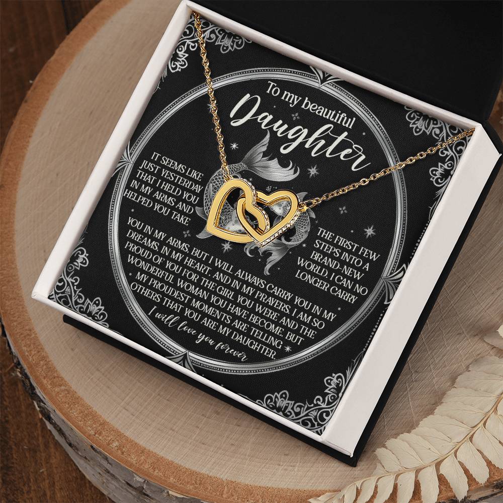 The Daughter-In My Prayers - Interlocking Hearts Necklace, with a rose gold finish, comes in a gift box featuring a heartfelt message for your daughter. Cubic zirconia crystals sparkle beside dried leaves on the wooden surface, symbolizing love and support.
