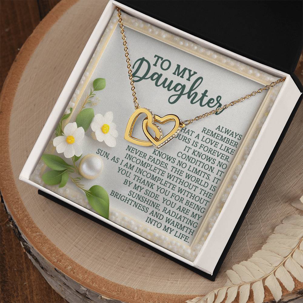 The Daughter-Love Forever - Interlocking Hearts Necklace, with a rose gold finish and cubic zirconia crystals, is elegantly displayed in a black box on a wooden surface. It includes a heartfelt message for your daughter on an elegant card, enhanced by white flowers.