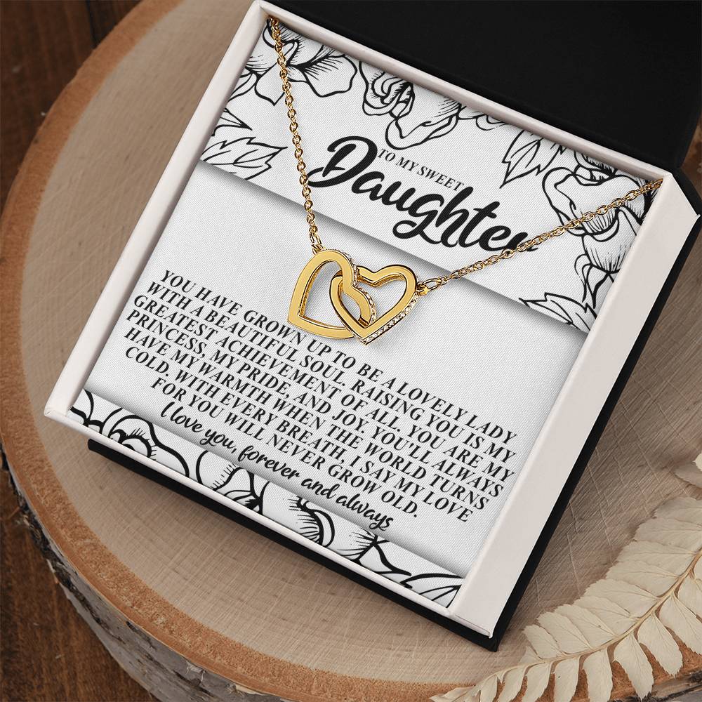 A close-up of the "Daughter-Beautiful Soul - Interlocking Hearts Necklace" in a jewelry box labeled "To My Sweet Daughter." Made from surgical steel, this exquisite piece features cubic zirconia crystals. The box's wooden surface and floral patterns amplify its loving message.