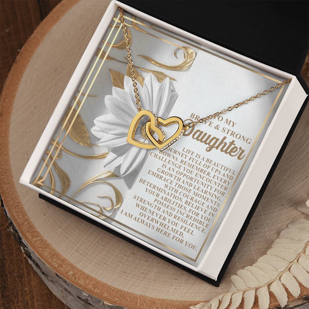 The Daughter-Here For You - Interlocking Hearts Necklace showcases a rose gold finish and cubic zirconia crystals in an open box. A white flower and inspirational message enhance the background, with the box placed on wood next to delicate dried white leaves.