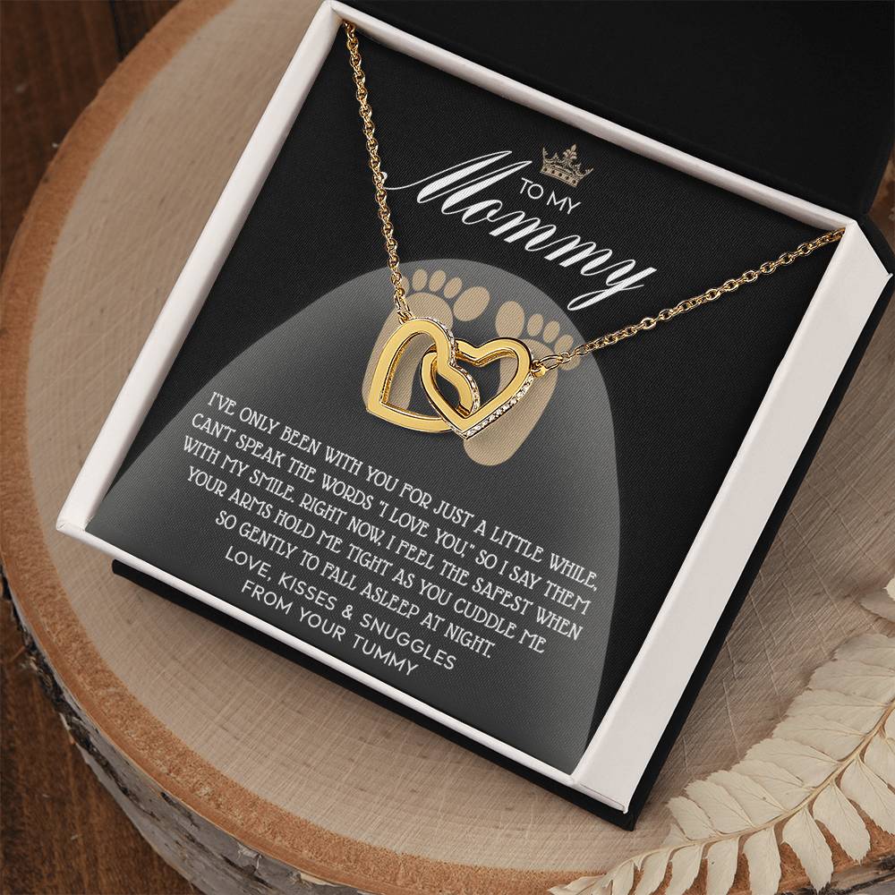 A rose gold "To Mama To Be, I Love You" Interlocking Hearts necklace adorned with cubic zirconia crystals rests on a black cushion, accompanied by a card reading "To My Mammy," featuring a sentimental message with a baby footprint design.