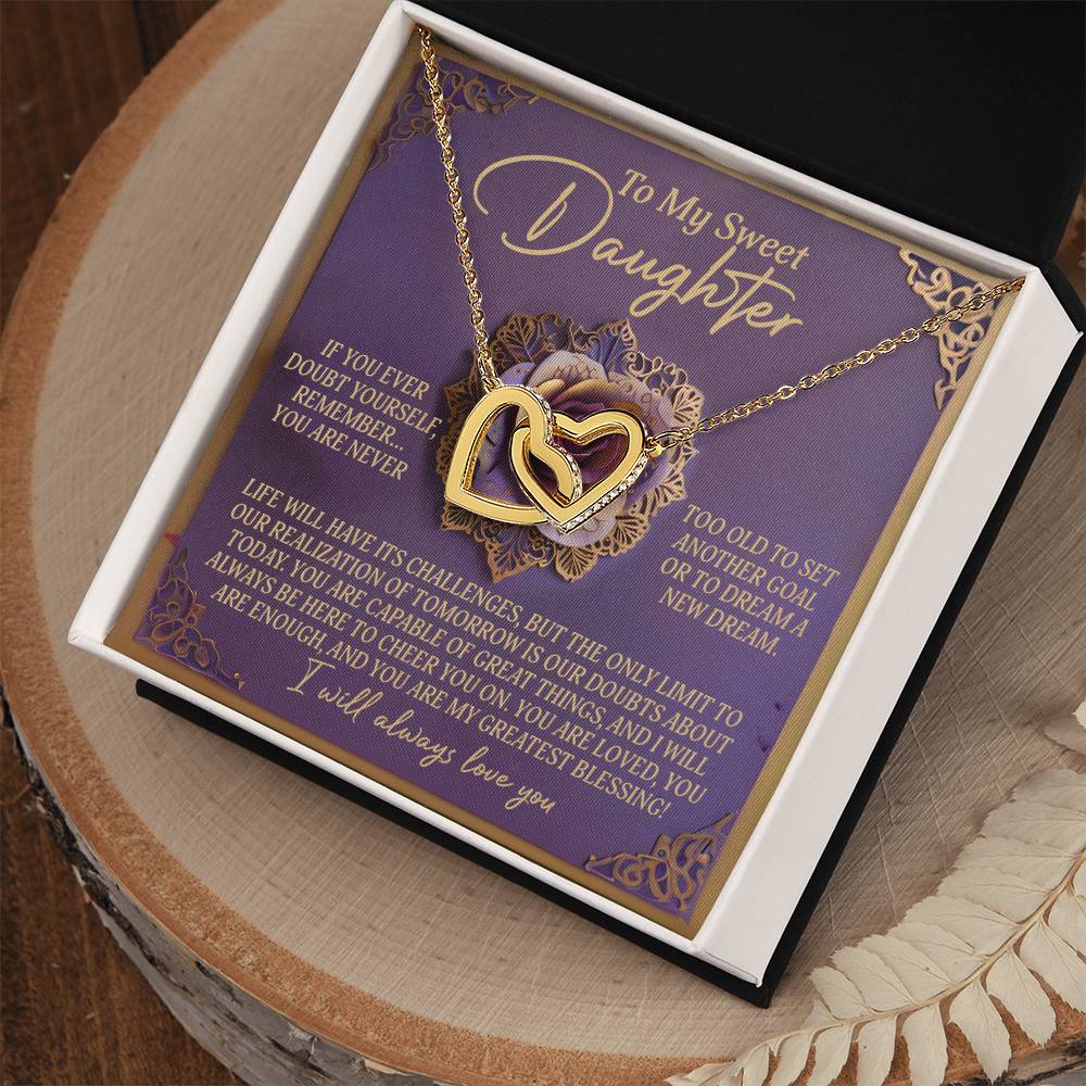 The Daughter-Greatest Blessing Interlocking Hearts Necklace, made of surgical steel with a rose gold finish and adorned with cubic zirconia crystals, is elegantly displayed on a purple card with floral designs and gold accents. The open box rests beautifully on a wooden surface.