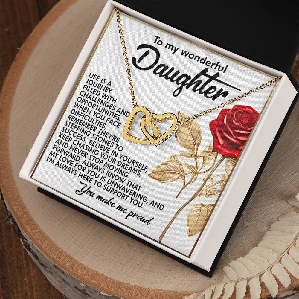 A heartfelt message for a daughter accompanies the Daughter-Journey Challenges - Interlocking Hearts Necklace, featuring cubic zirconia crystals and a red rose illustration. The open rose gold-finished box elegantly rests on a wooden surface.