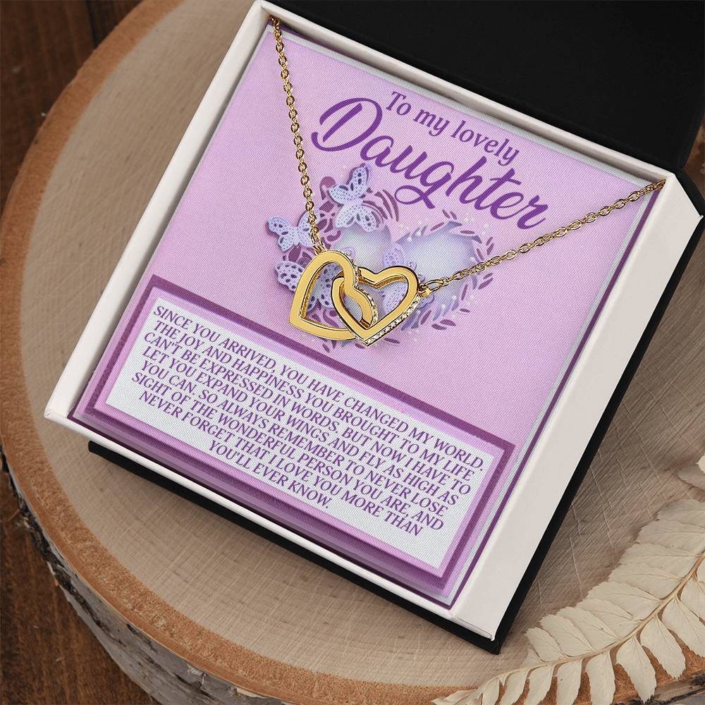 The Daughter-Expand Wings Interlocking Hearts Necklace features a rose gold finish and cubic zirconia crystals, placed on a purple card with "To my lovely Daughter" and a heartfelt message, elegantly boxed in black and white on a wooden surface.