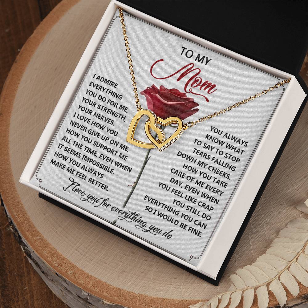 The Mom-Be Fine - Interlocking Hearts Necklace comes in a black box and features a heart-shaped pendant with rose gold finish, adorned with cubic zirconia crystals. The lid displays an emotional message of gratitude to a mother over a red rose background.