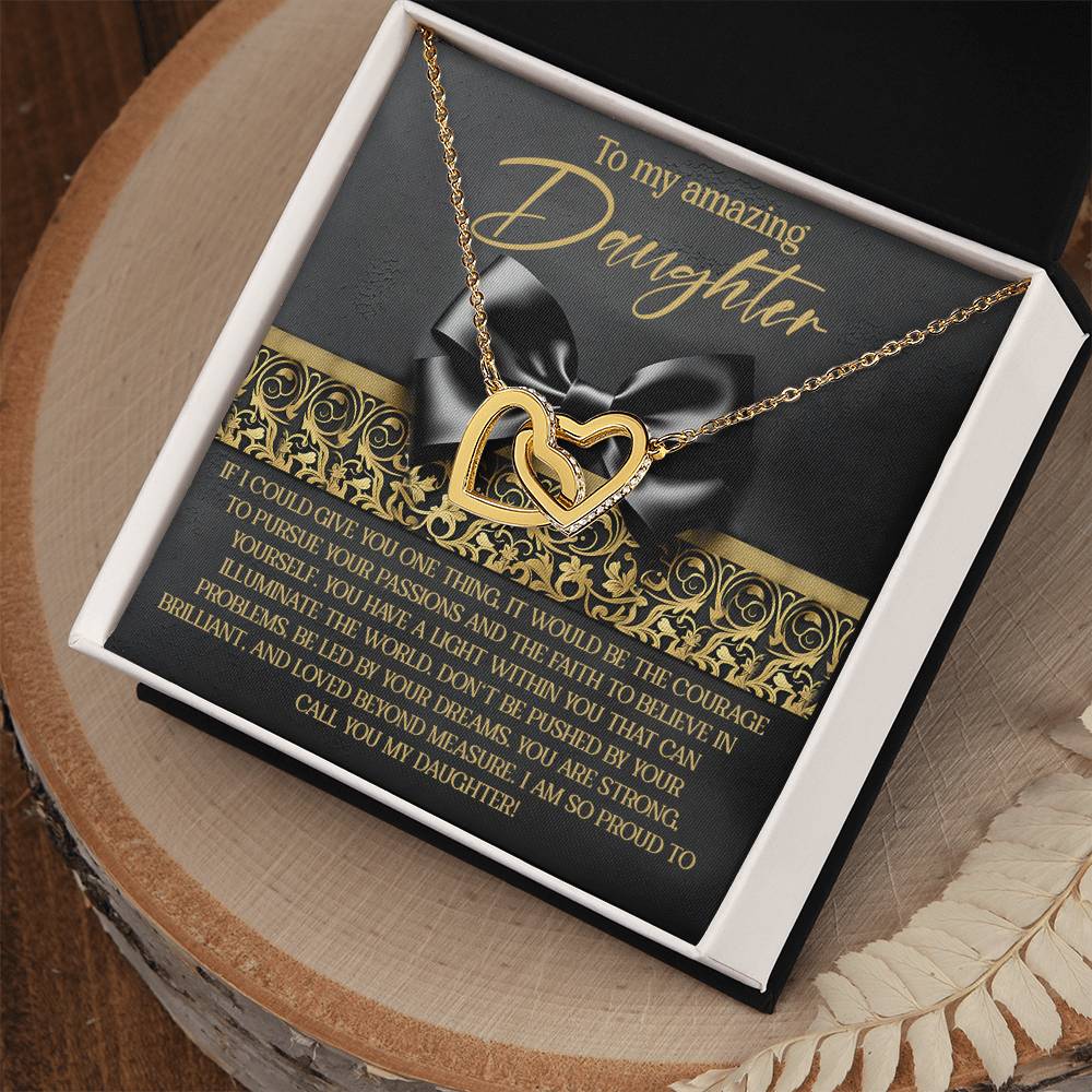The Daughter-Pursue Passions - Interlocking Hearts Necklace, adorned with cubic zirconia crystals and a rose gold finish, comes in an ornate open box. The liner reads "To my amazing daughter" with an emotional message. The box rests gracefully on a wooden surface.