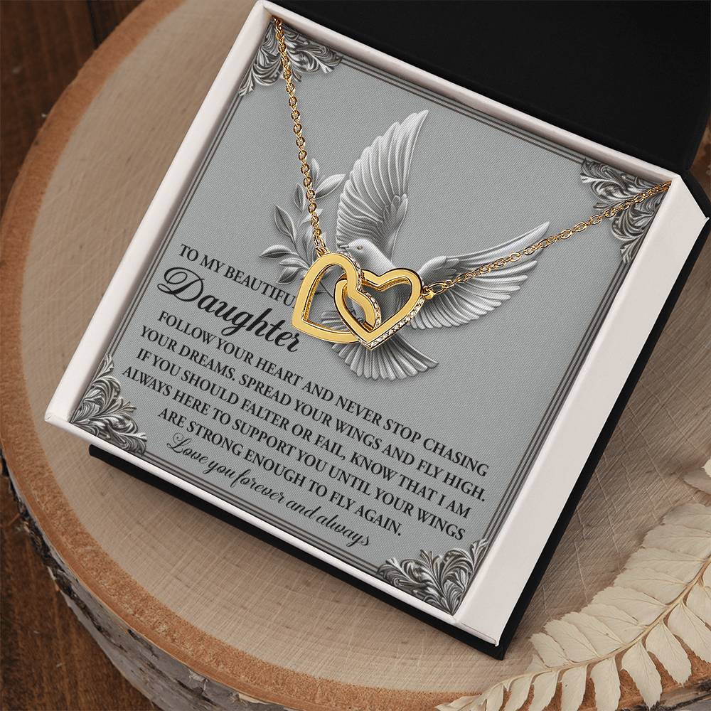 The Daughter-Your Wings Interlocking Hearts Necklace, featuring cubic zirconia and an 18k yellow gold finish, sits in an open box with a dove illustration backdrop and a heartfelt message for a daughter.