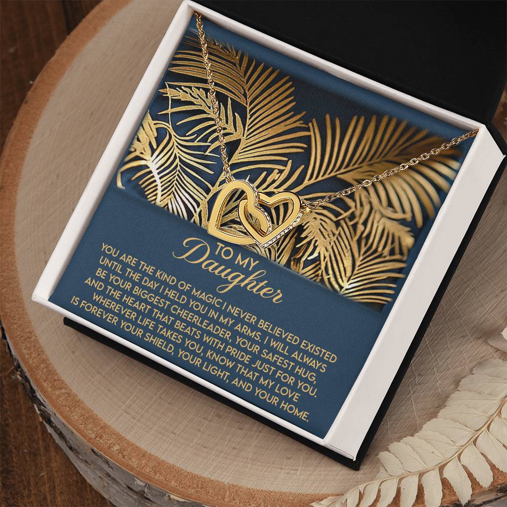 The "Daughter-Your Home - Interlocking Hearts Necklace" with a rose gold finish rests in an open jewelry box, featuring a heartfelt message of love and protection to a daughter, set against a decorative backdrop of gold palm leaves.