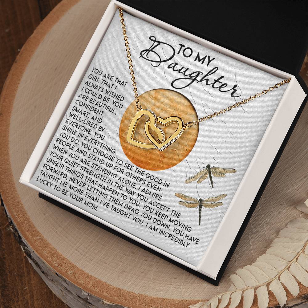 The "Daughter-Quiet Strength - Interlocking Hearts Necklace" comes in a gift box featuring dragonfly illustrations and an emotional message titled "To My Daughter." The necklace is adorned with cubic zirconia crystals, and the wooden background is accented with decorative leaves.