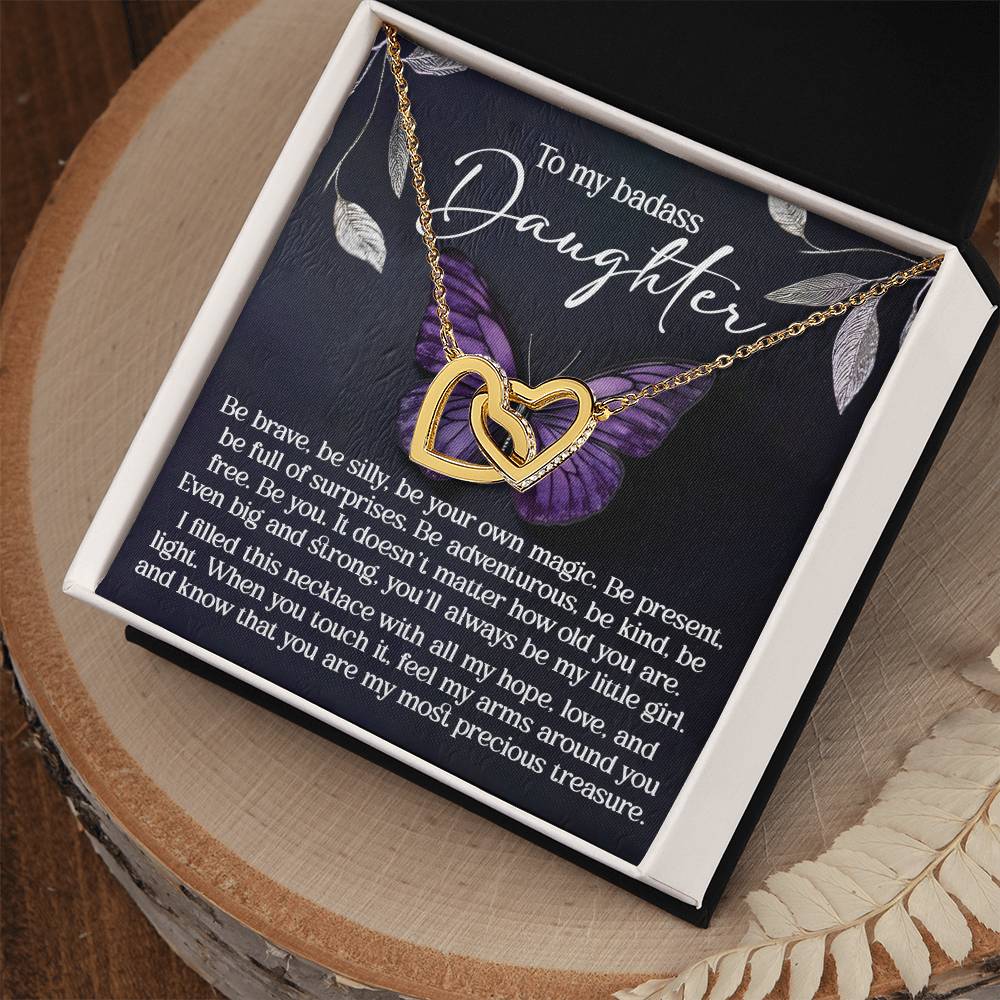 The Daughter-Big And Strong Interlocking Hearts necklace, featuring a rose gold finish and cubic zirconia crystals, rests in a gift box. With a purple butterfly backdrop, the message inside honors your daughter's bravery, magic, and love, calling her "a most precious treasure.