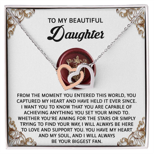 The Daughter-Biggest Fan Interlocking Hearts Necklace features a heart-shaped pendant with a rose gold finish and sparkling cubic zirconia crystals, presented on an ornate card with a loving message to a daughter.