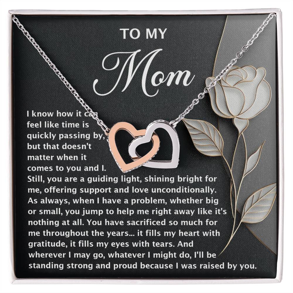 The Mom-Guiding Light - Interlocking Hearts Necklace, adorned with cubic zirconia crystals and a rose gold finish, is presented on a card with a heartfelt message for Mom. The dark background and decorative rose emphasize gratitude, love, guidance, and sacrifice.
