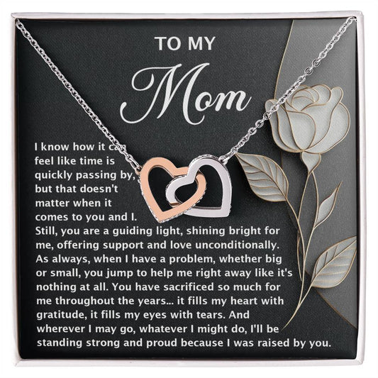 The Mom-Guiding Light - Interlocking Hearts Necklace, adorned with cubic zirconia crystals and a rose gold finish, is presented on a card with a heartfelt message for Mom. The dark background and decorative rose emphasize gratitude, love, guidance, and sacrifice.
