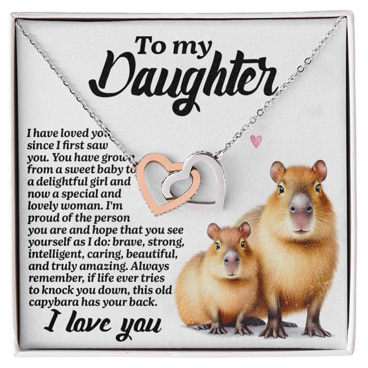 The Daughter-Sweet Baby Interlocking Hearts Necklace, with a rose gold finish and cubic zirconia crystals, is beautifully presented on a card featuring two capybaras and a heartfelt "To my Daughter" message expressing love and admiration.