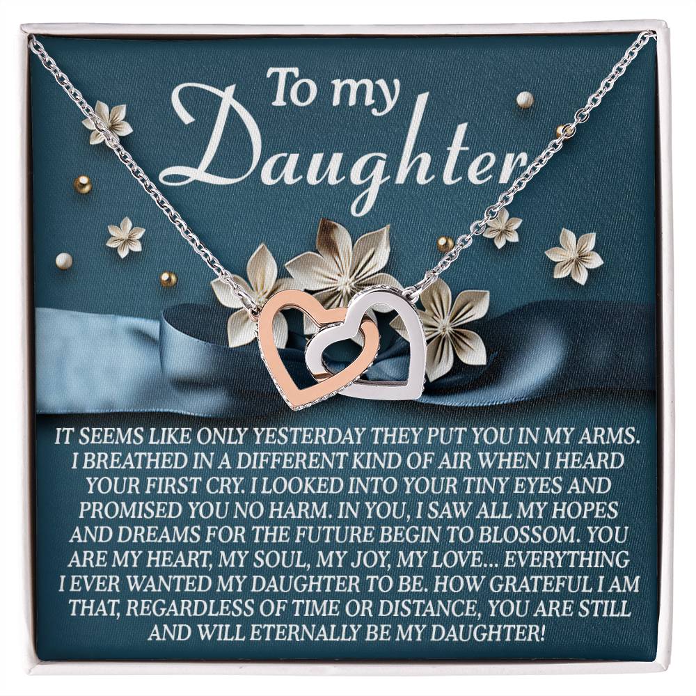 The Daughter-Hopes Dreams Interlocking Hearts Necklace with cubic zirconia crystals is beautifully displayed in a jewelry gift box. The lid reads "To my Daughter" alongside a heartfelt message, while flowers in the background add elegance, making it the ideal gift for her.