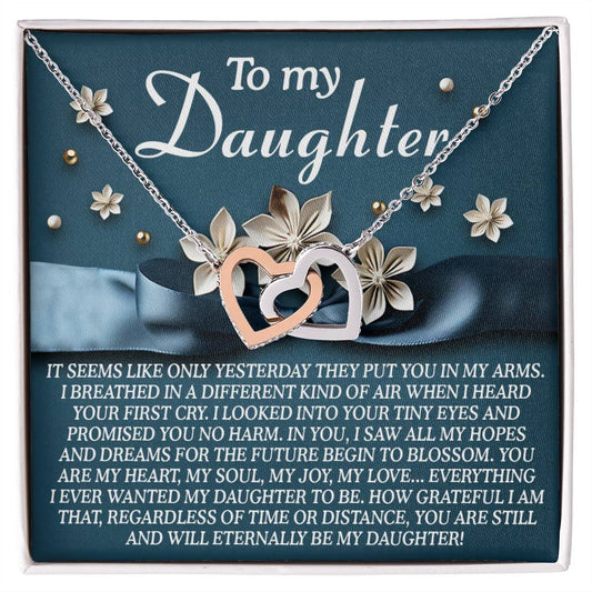The Daughter-Hopes Dreams Interlocking Hearts Necklace with cubic zirconia crystals is beautifully displayed in a jewelry gift box. The lid reads "To my Daughter" alongside a heartfelt message, while flowers in the background add elegance, making it the ideal gift for her.