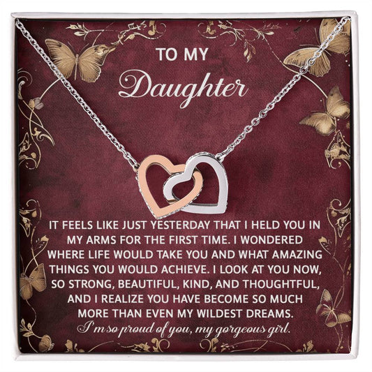 The Daughter-Wildest Dreams - Interlocking Hearts Necklace, with cubic zirconia crystals and a rose gold finish, is displayed on a decorative card. This card features butterfly and floral motifs and conveys a heartfelt message expressing pride and love for a daughter.