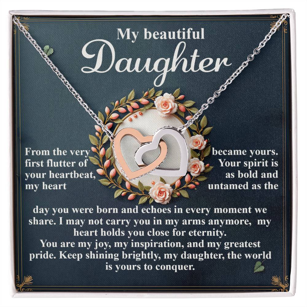 The Daughter-First Flutter Interlocking Hearts Necklace, adorned with cubic zirconia and a rose gold finish, rests on a box saying "My beautiful Daughter." The backdrop conveys love and pride amidst fluttering heartbeats and an eternal spirit, all encircled by pink roses in a floral wreath.