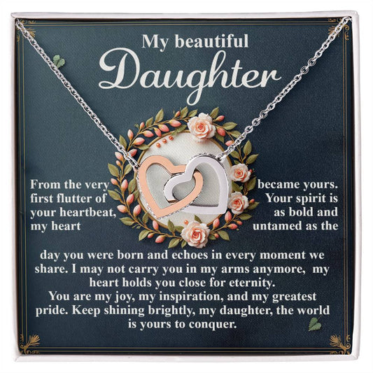 The Daughter-First Flutter Interlocking Hearts Necklace, adorned with cubic zirconia and a rose gold finish, rests on a box saying "My beautiful Daughter." The backdrop conveys love and pride amidst fluttering heartbeats and an eternal spirit, all encircled by pink roses in a floral wreath.