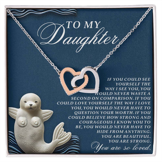 The Daughter-So Loved Interlocking Hearts Necklace features a rose gold and silver heart adorned with cubic zirconia crystals. It comes on a dark blue card for a daughter, featuring a white illustration of a smiling seal holding a heart.