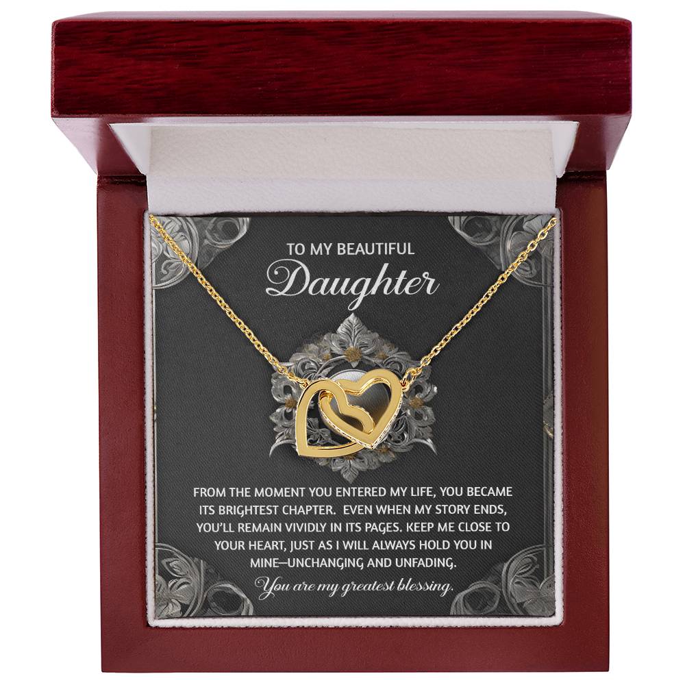 The Daughter-Greatest Blessing - Interlocking Hearts Necklace features a gold heart design with cubic zirconia crystals, set in a box with a heartfelt message about love and memories. Its surgical steel and rose gold finish complement the decorative background.